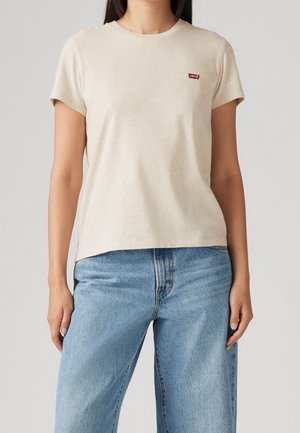 Femme portant un t-shirt beige à manches courtes avec un petit logo rouge Levi's, associé à un jean taille haute bleu clair.