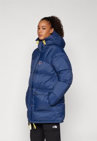 Marine blauwe pufferjack met een hoge kraag, capuchon en gele trekkoorden. Voorzien van drukknoopzakken en een logo-label op de borst.