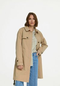 Beige trenchcoat met een dubbelzijdig ontwerp, knoopaccenten en lange mouwen. Graagt over een gestreept shirt, gecombineerd met lichtblauwe spijkerbroek.