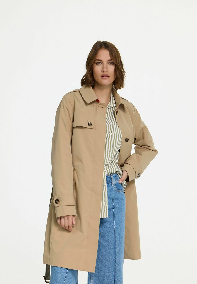 Beige trenchcoat met een dubbelzijdig ontwerp, knoopaccenten en lange mouwen. Graagt over een gestreept shirt, gecombineerd met lichtblauwe spijkerbroek.