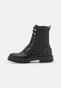 Stivale da combattimento in pelle nera con suola spessa, occhielli dorati, cerniera laterale e motivo geometrico stampato sul pannello alla caviglia.