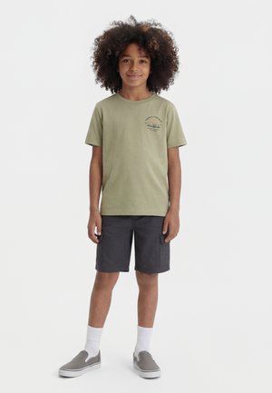 Enfant aux cheveux bouclés portant un t-shirt graphique vert olive, un short cargo anthracite, des chaussettes blanches et des chaussures grises sans lacets, debout et souriant.