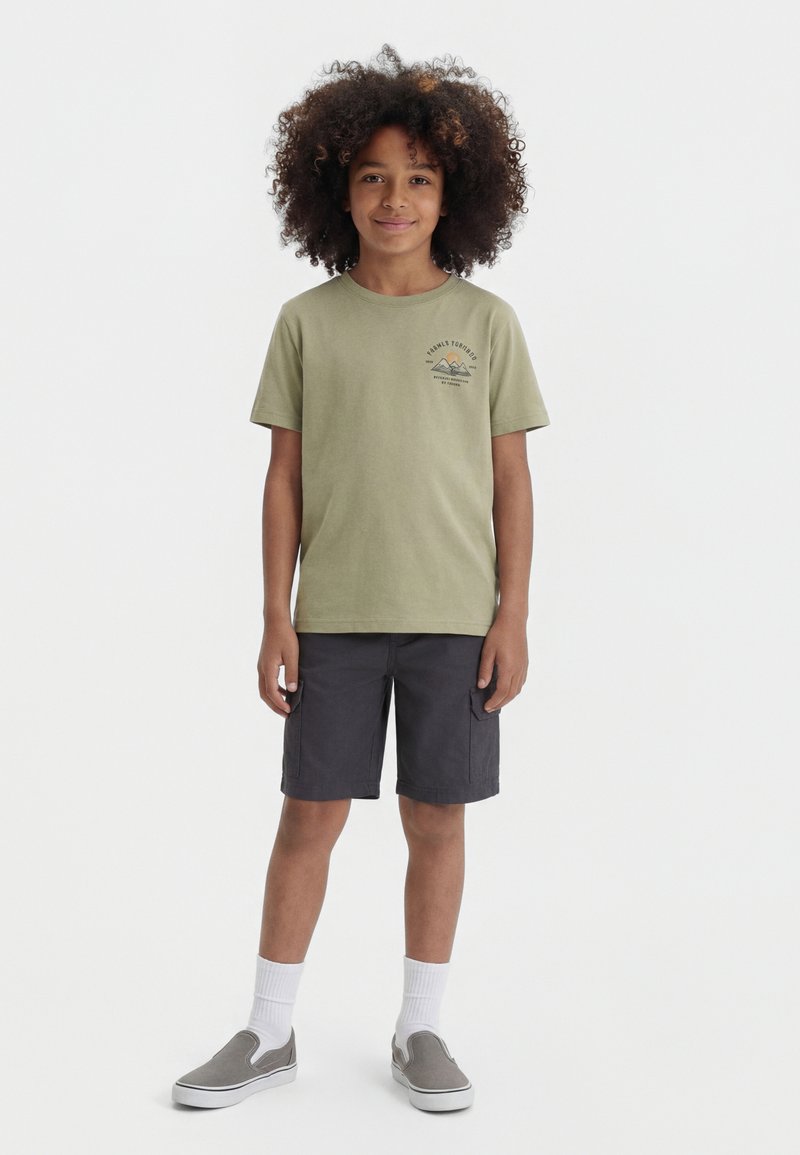 Enfant aux cheveux bouclés portant un t-shirt graphique vert olive, un short cargo anthracite, des chaussettes blanches et des chaussures grises sans lacets, debout et souriant.