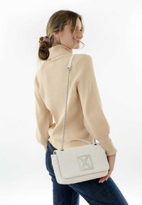 Borsa a tracolla bianca trapuntata con una placca argento e catena. Il modello indossa un dolcevita a coste beige chiaro e jeans scuri.
