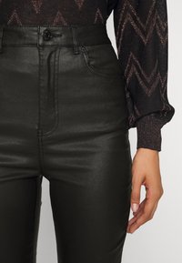 Kvinna som bär högmidjade svarta läderbyxor och en långärmad svart topp med metalliska zigzag-mönster, står med höger hand avslappnad.