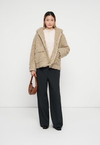Giacca in pelliccia sintetica beige con colletto ampio e due tasche frontali, indossata sopra una blusa chiara e abbinata a pantaloni a righe sottili e una borsa marrone.