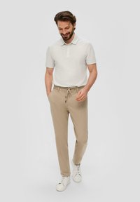 Beige Baumwollmischungshose mit verstellbarem Kordelzugbund, kombiniert mit einem hellgrauen Poloshirt. Weiße Sneakers runden das Outfit ab.