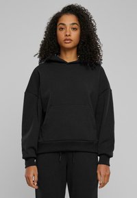 Felpa nera con cappuccio, dal taglio morbido, grande tasca frontale, polsini e orlo a costine, texture morbida, indossata con pantaloni abbinati.