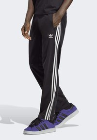 Pantalones deportivos negros con diseño de tres rayas blancas, bolsillos laterales y textura suave. Llevados con zapatillas de ante púrpuras con detalles en negro.