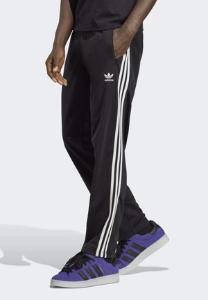 Pantaloni sportivi neri con design a tre strisce bianche, tasche laterali e texture morbida. Indossati con sneakers in camoscio viola con accenti neri.