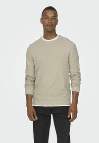 Un pull beige clair avec un motif en losanges texturé, présentant une encolure ronde à côtes et des poignets côtelés. Porté par-dessus un jean foncé.