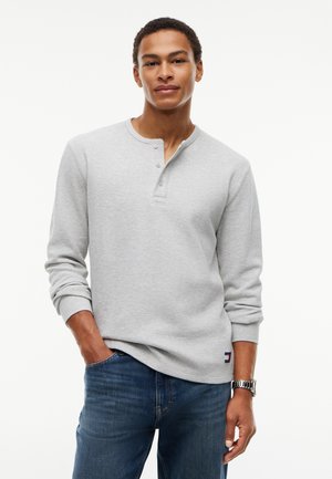 Ung mand iført en lys grå langærmet henley-shirt og blå jeans, med den ene hånd i lommen, stående mod en hvid baggrund.