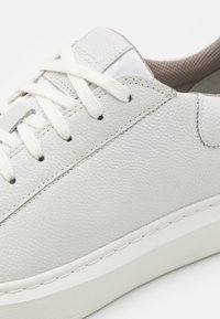 White leather sneaker featuring a textured surface, flat white laces, subtle stitching, and a contrasting beige interior. Smooth rubber sole.