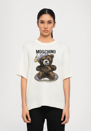 Vit oversized stickad t-shirt med en björngrafik i brunt, med en design av en blå och vit kopp och tallrik ovanför. Moschino-logotyp inkluderad.