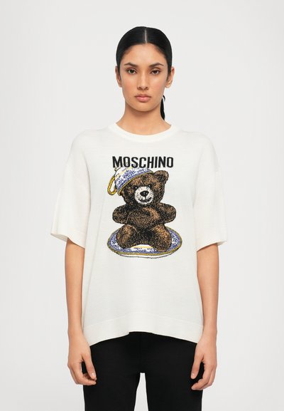 T-shirt en tricot surdimensionné blanc avec un graphique d'ours marron, accompagné d'un design de tasse et d'assiette bleu et blanc au-dessus. Marque Moschino incluse.