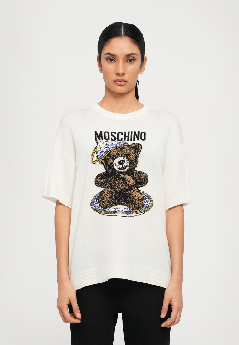 T-shirt bianco oversize in maglia con grafica di un orso marrone, decorata con un design di tazza e piatto blu e bianco sopra. Include il marchio Moschino.