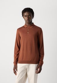 Mann mit kurzen lockigen Haaren, der ein braunes Langarm-Polohemd und hellbeige Hose trägt, vor einem schlichten hellen Hintergrund stehend.