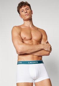 Bijeli pamučni bokserice s tirkiznim elastičnim pojasom na kojem je otisnut "LACOSTE" logo. Uski dizajn ističe konture nogu i struka.
