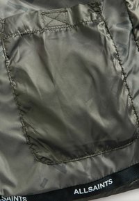 Primo piano di un tessuto verde oliva con una toppa rettangolare cucita e il marchio "ALLSAINTS" in bianco sull'orlo nero.