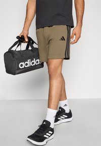 Melna Adidas sporta soma, olīvu zaļas šortus ar trim melnām svītrām, melnas sporta kurpes ar baltām detaļām un baltas zeķes.