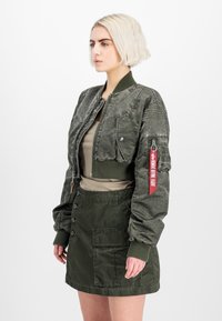 Olijfgroene bomberjack met een cropped ontwerp, ritssluiting, rib gebreide manchetten en een rode labeldetail op de mouw. Gladde, textuurstof.