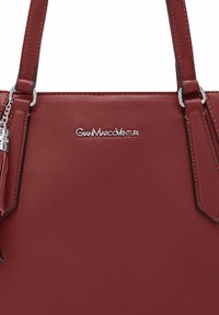 Bolso de piel roja con textura suave, herrajes plateados y logo exhibido de manera prominente. Cuenta con doble asa y un borlón lateral.