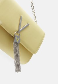 Gul läderhandväska med silverdetaljer, prydd med en 'V' logotyp och metallkedjetassel. Slät textur och stadig design.