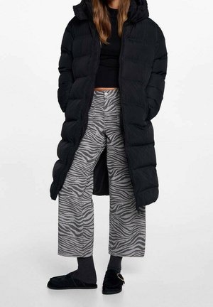 Femme portant un long manteau matelassé noir, un haut noir court, un pantalon ample à larges jambes gris à rayures zèbre, des chaussettes foncées et des chaussons noirs avec boucle.