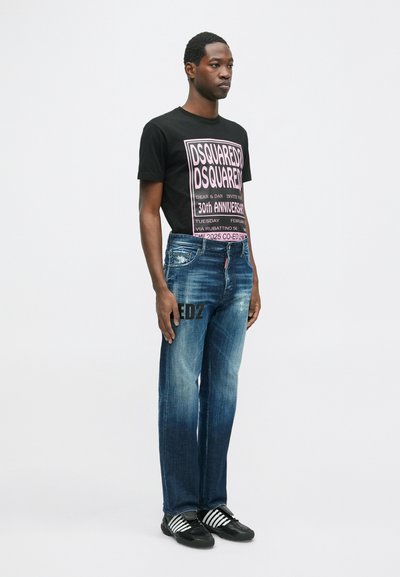 Čierne grafické tričko s ružovým textom, modré vyblednuté džínsy s potlačou "ED2" a čierne topánky s bielymi pruhmi, stojaci pozícia na svetlom pozadí.