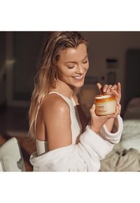 En klar glasburk med kroppskräm och ett gult lock, hålls av en person i vit morgonrock, som visar en slät, krämig konsistens.