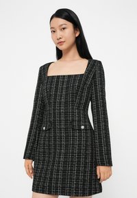 Rochie din tweed cu carouri alb-negru, cu decolteu pătrat, mâneci lungi și două buzunare frontale ce au nasturi asemănători perlelor.
