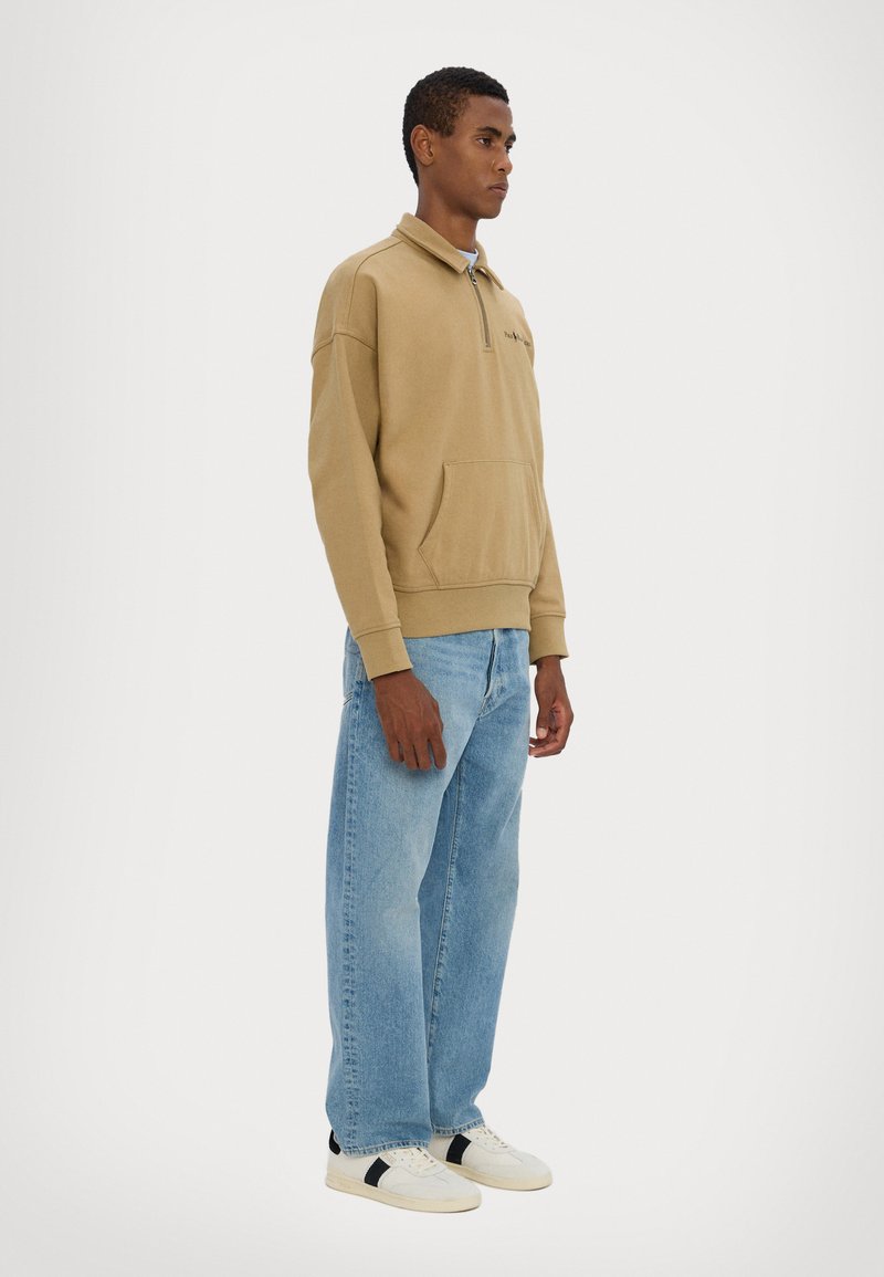 Tan zip-up sweatshirt with a front pocket, paired with light blue loose-fitting jeans and white sneakers featuring black accents.