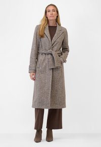 Brun, vävd ullblandning kappa med bälte, slagkrage och två framfickor, bärs över en mörkbrun topp och vida byxor.