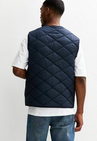 Man som bär en marinblå vadderad väst över en vit T-shirt och blå jeans, vänd bort från en ren vit bakgrund.