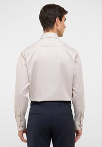 Camicia a maniche lunghe color beige chiaro con un motivo testurizzato, polsini con bottoni e colletto classico, indossata con pantaloni blu scuro.