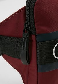 Rode stoffen tas met zwarte accenten. Voorzien van een ritsluiting, webbing schouderband en cirkelvormig hardware detail. Getextureerd oppervlak en stevige constructie.