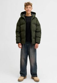 Giubbotto imbottito verde scuro, con cappuccio, chiusura lampo e trama trapuntata. Abbinato a jeans blu dal taglio comodo e scarpe beige.