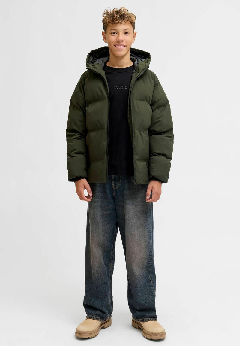 Giubbotto imbottito verde scuro, con cappuccio, chiusura lampo e trama trapuntata. Abbinato a jeans blu dal taglio comodo e scarpe beige.