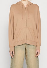 Beige zip-up hoodie with a hood, front pockets, and ribbed cuffs. Made from soft fabric with a smooth texture and no patterns.