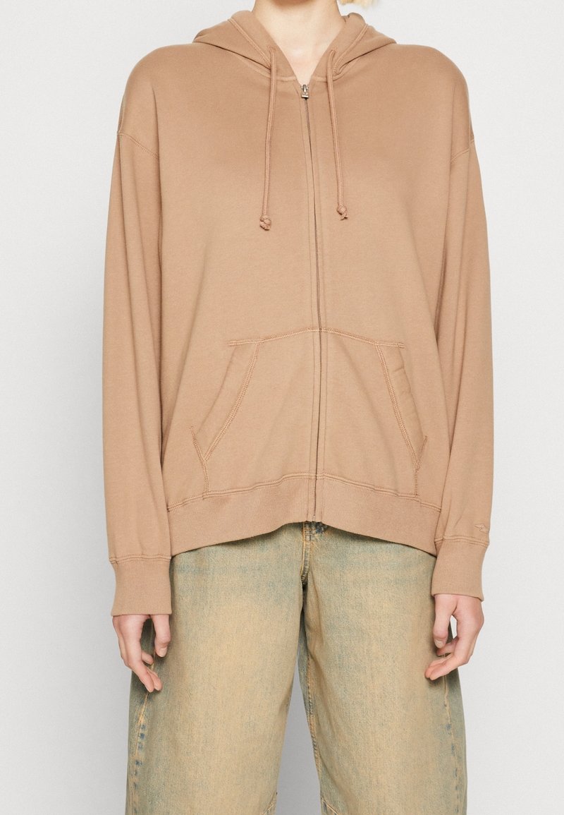 Beige zip-up hoodie with a hood, front pockets, and ribbed cuffs. Made from soft fabric with a smooth texture and no patterns.