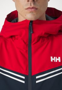Rote Kapuzenjacke mit einem navyfarbenen unteren Bereich, ausgestattet mit weißen Streifen und einem Logo. Der Stoff wirkt glatt und hat einen Reißverschluss.