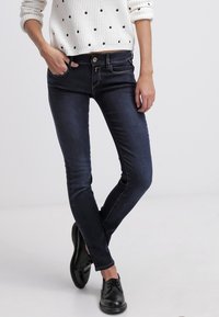 Jeans skinny azul oscuro de mezclilla; cuenta con un cierre frontal con botón y dos bolsillos frontales. Combinado con un suéter blanco con lunares.