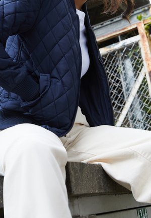 Personne portant une veste matelassée bleu marine et un pantalon blanc assise sur un rebord en béton près d'une clôture en grillage.