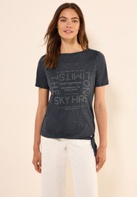 Camiseta de manga corta azul marino con tejido texturizado y texto impreso. Estilo con un detalle de nudo en el lado. Combinada con pantalones blancos.