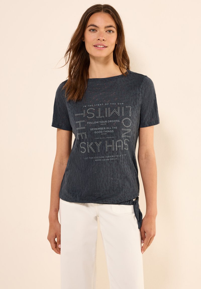 Camiseta de manga corta azul marino con tejido texturizado y texto impreso. Estilo con un detalle de nudo en el lado. Combinada con pantalones blancos.