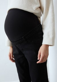 Mustus denimist emade püksid veniva musta vöökoha ja kreemja kampsuni all, rõhutades ümmargust kõhtu.