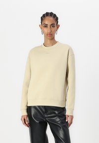 Sudadera de cuello redondo beige con puños y dobladillo acanalados. Tejido suave con un mínimo de marca. Combinada con pantalones de imitación cuero negros.