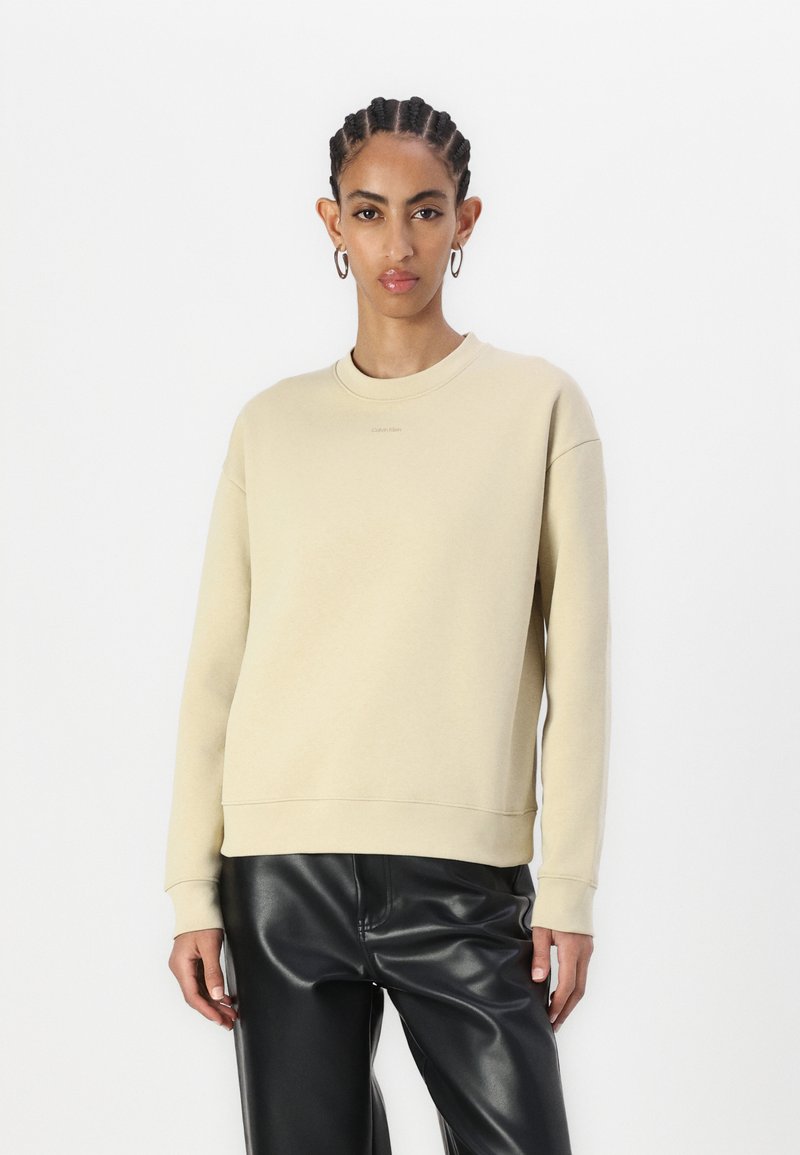 Sudadera de cuello redondo beige con puños y dobladillo acanalados. Tejido suave con un mínimo de marca. Combinada con pantalones de imitación cuero negros.