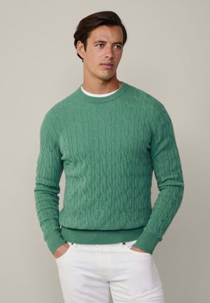 Maglione a girocollo lavorato a maglia in verde chiaro. Presenta polsini e orlo a costine. Indossato sopra una camicia bianca, abbinato a pantaloni bianchi.