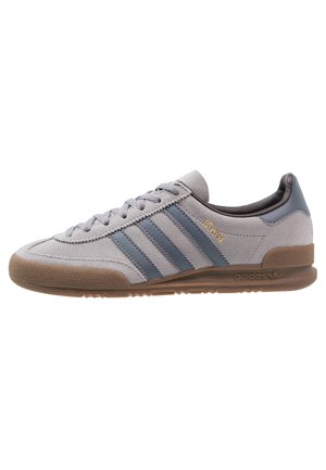 Szary zamszowy sneaker Adidas z niebieskimi paskami, gumową podeszwą i złotym napisem „Jeans” z boku.